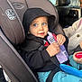 toddler, child, car_seat, hoodie, snack_pouch, blue_eyes, seatbelt, car_interior, plush_toy, clothing, person, seat, window, vehicle, sitting, black_hoodie, blue_pants, hand, face, curious