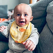 Bodi is registered to the contest to win money with this photo: baby, bib, bright_eyes, child, closeup, couch, cozy, cute, duck, gray, happy, home, indoor, infant, knitted_clothing, person, portrait, reaching, sitting, smiling