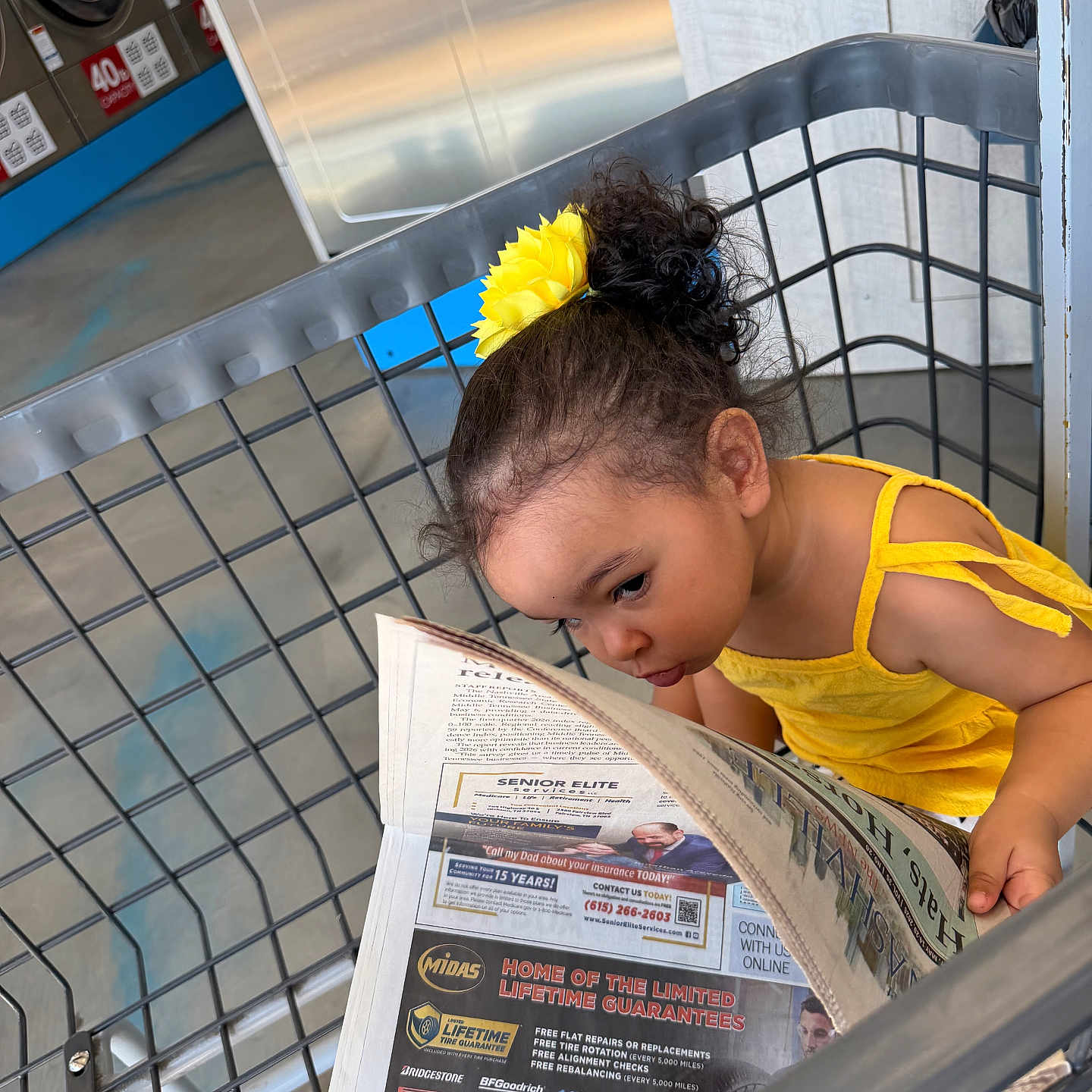 Yamanie is registered to the contest to win money with this photo: child, girl, shopping_cart, newspaper, yellow_dress, hair_accessory, laundromat, curly_hair, reading, metal, floor, background, clothing, indoor, person, portrait, casual, cute, concentration, toddler