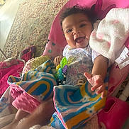 Delilah is registered to the contest to win money with this photo: baby, smiling, infant, blanket, colorful, toy, indoor, happy, child, seat, curly_hair, patterned_clothing, carpet, home, reaching_hand, cozy, playful, cute, toddler, baby_bouncer