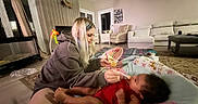 Delilah is registered to the contest to win money with this photo: baby, child, feeding, woman, indoor, living_room, couch, armchair, blanket, play_mat, toy, care, feeding_spoon, female, person, floor, curtains, hair, clothing, comfort