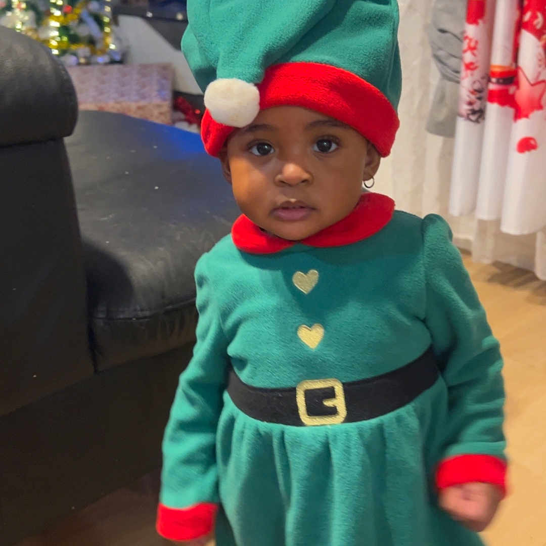 Grace-Edah a rejoint le concours — aidez-le/la à gagner de superbes lots ! child, christmas_tree, curious_expression, curtains, earrings, elf_costume, festive, green_dress, hat, holiday, holiday_decor, indoor, person, portrait, red_trim, socks, sofa, television, toddler, wooden_floor