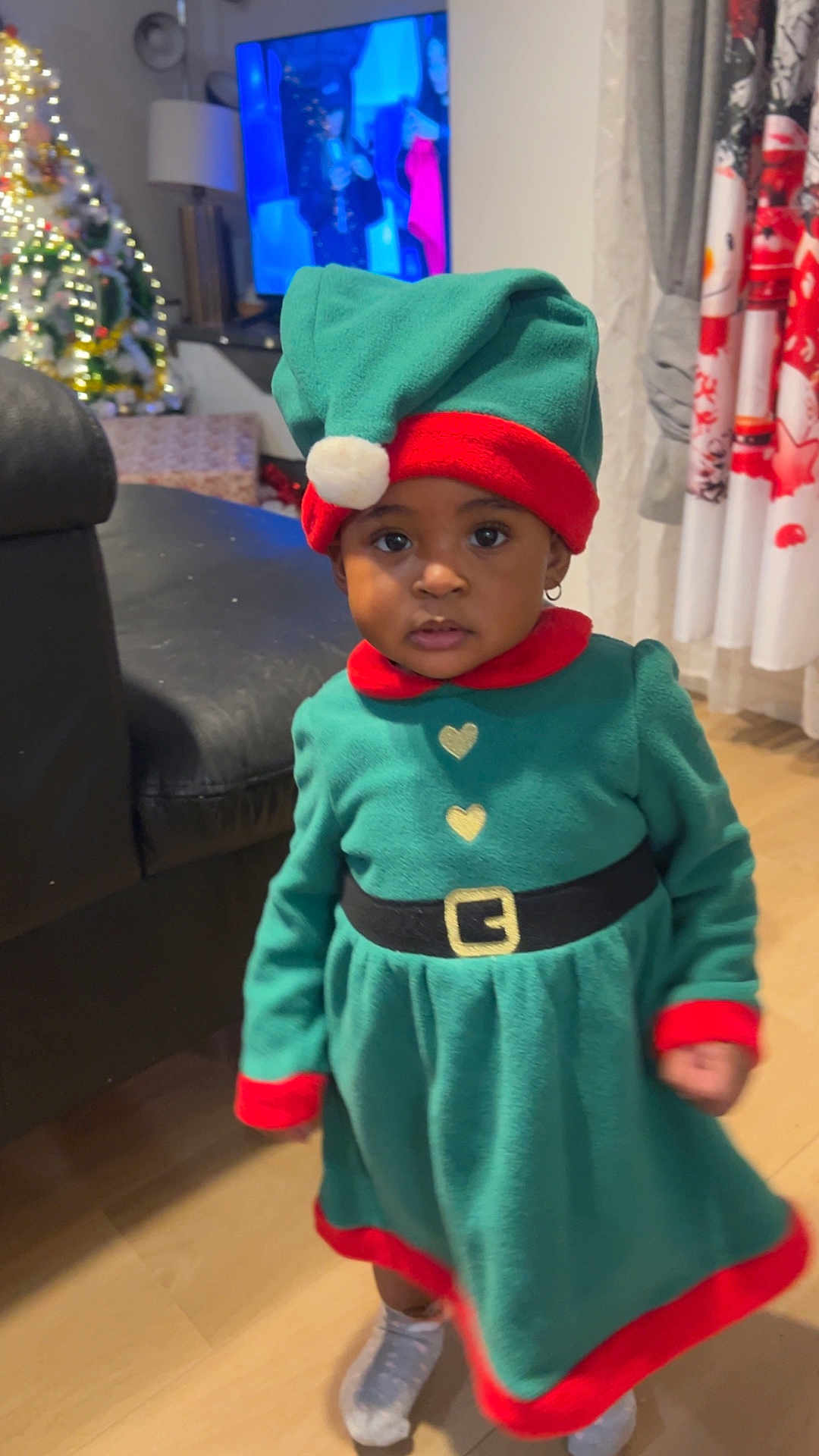 Grace-Edah a rejoint le concours — aidez-le/la à gagner de superbes lots ! toddler, child, elf_costume, green_dress, red_trim, holiday, christmas_tree, indoor, curious_expression, hat, socks, wooden_floor, sofa, curtains, earrings, television, festive, holiday_decor, person, portrait
