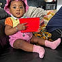 toddler, child, pink_hat, pink_socks, red_tablet, bracelet, couch, living_room, indoor, curious, clothing, person, furniture, technology, baby, earrings, sitting, portrait, black_leather, home