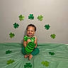 toddler, child, smiling, green_bowtie, green_pants, bare_chest, shamrocks, st_patricks_day, green_cup, floor_covering, decorations, celebration, happy, indoor, curly_hair, cute, sitting, tooth_grin, festive, party
