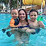child, toddler, woman, pool, water_slide, swimming_pool, water, smiling, family, goggles, swimwear, swimsuit, outdoors, palm_trees, reflection, wet_hair, vacation, playful, laughter, lounge_chairs