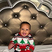 Makenai joined the competition — help win amazing prizes! baby, child, smiling, sweater, christmas, holiday, bed, remote_control, happy, indoors, tufted_headboard, furniture, person, portrait, cute, winter_clothing, sitting, joyful, home, cozy