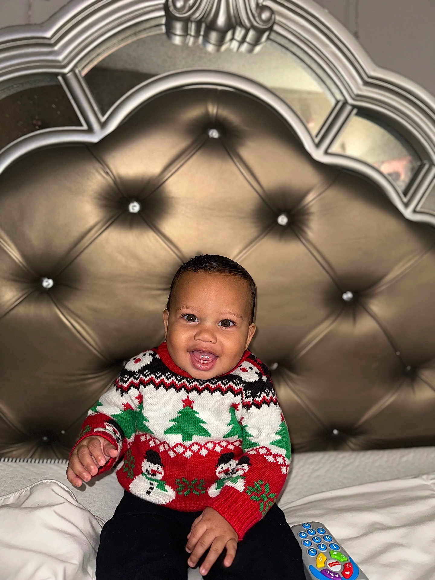 Makenai joined the competition — help win amazing prizes! baby, child, smiling, sweater, christmas, holiday, bed, remote_control, happy, indoors, tufted_headboard, furniture, person, portrait, cute, winter_clothing, sitting, joyful, home, cozy