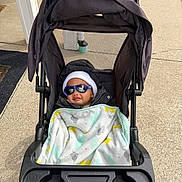 Makenai joined the competition — help win amazing prizes! baby, stroller, blanket, sunglasses, hat, jacket, outside, sidewalk, person, child, cute, warm_clothing, baby_accessory, sun_protection, urban, daytime, comfort, relaxing, infant, outdoor
