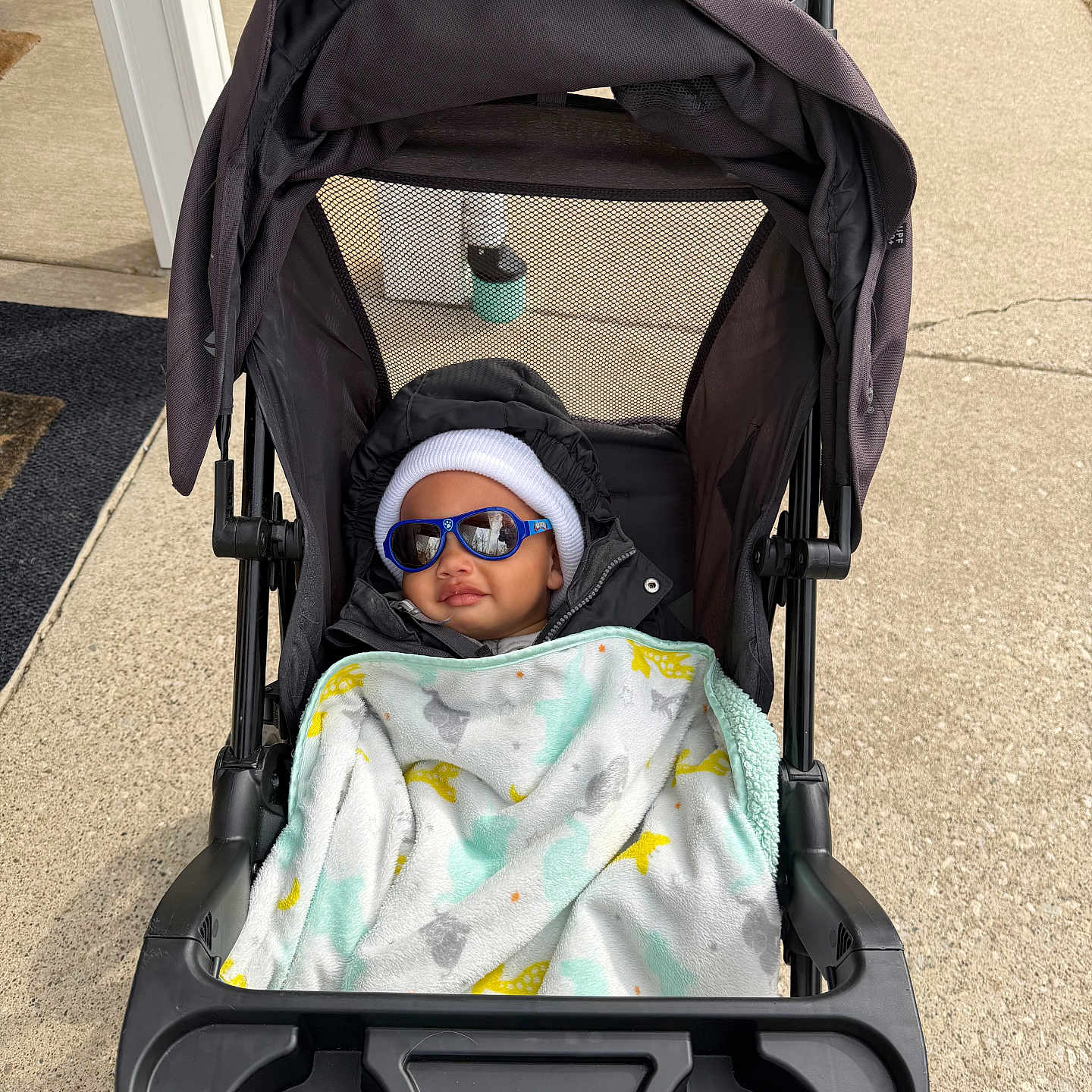 Makenai joined the competition — help win amazing prizes! baby, stroller, blanket, sunglasses, hat, jacket, outside, sidewalk, person, child, cute, warm_clothing, baby_accessory, sun_protection, urban, daytime, comfort, relaxing, infant, outdoor