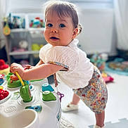 Mahée a rejoint le concours — aidez-le/la à gagner de superbes lots ! activity_table, baby_shoes, blurred_background, child, colorful, curious, cute, floor_mat, indoor, natural_light, person, playroom, short_hair, smiling, soft_light, standing, toddler, toy, white_clothing, window