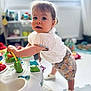 activity_table, baby_shoes, blurred_background, child, colorful, curious, cute, floor_mat, indoor, natural_light, person, playroom, short_hair, smiling, soft_light, standing, toddler, toy, white_clothing, window