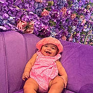 Lynae a rejoint le concours — aidez-le/la à gagner de superbes lots ! baby, smiling, pink_clothing, hat, purple_couch, flower_wall, indoor, happy, infant, cute, portrait, child, plush_furniture, floral_pattern, relaxing, toddler, bright_colors, sitting, skin, cheerful