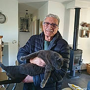 Iron a rejoint le concours — aidez-le/la à gagner de superbes lots ! elderly_man, glasses, gray_cat, holding, smiling, indoor, living_room, wood_stove, modern_decor, casual_clothing, puffer_jacket, furniture, cushion, wall_clock, plant, basket, floor_tiles, portrait, happy, relaxed_cat
