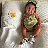 Kayani is registered to the contest to win money with this photo: baby, child, smiling, green_outfit, headband, white_blanket, sitting, cute, happy, infant, footwear, sandals, indoor, portrait, skin, curly_hair, sign, illustration, text, cozy