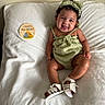baby, child, smiling, green_outfit, headband, white_blanket, sitting, cute, happy, infant, footwear, sandals, indoor, portrait, skin, curly_hair, sign, illustration, text, cozy