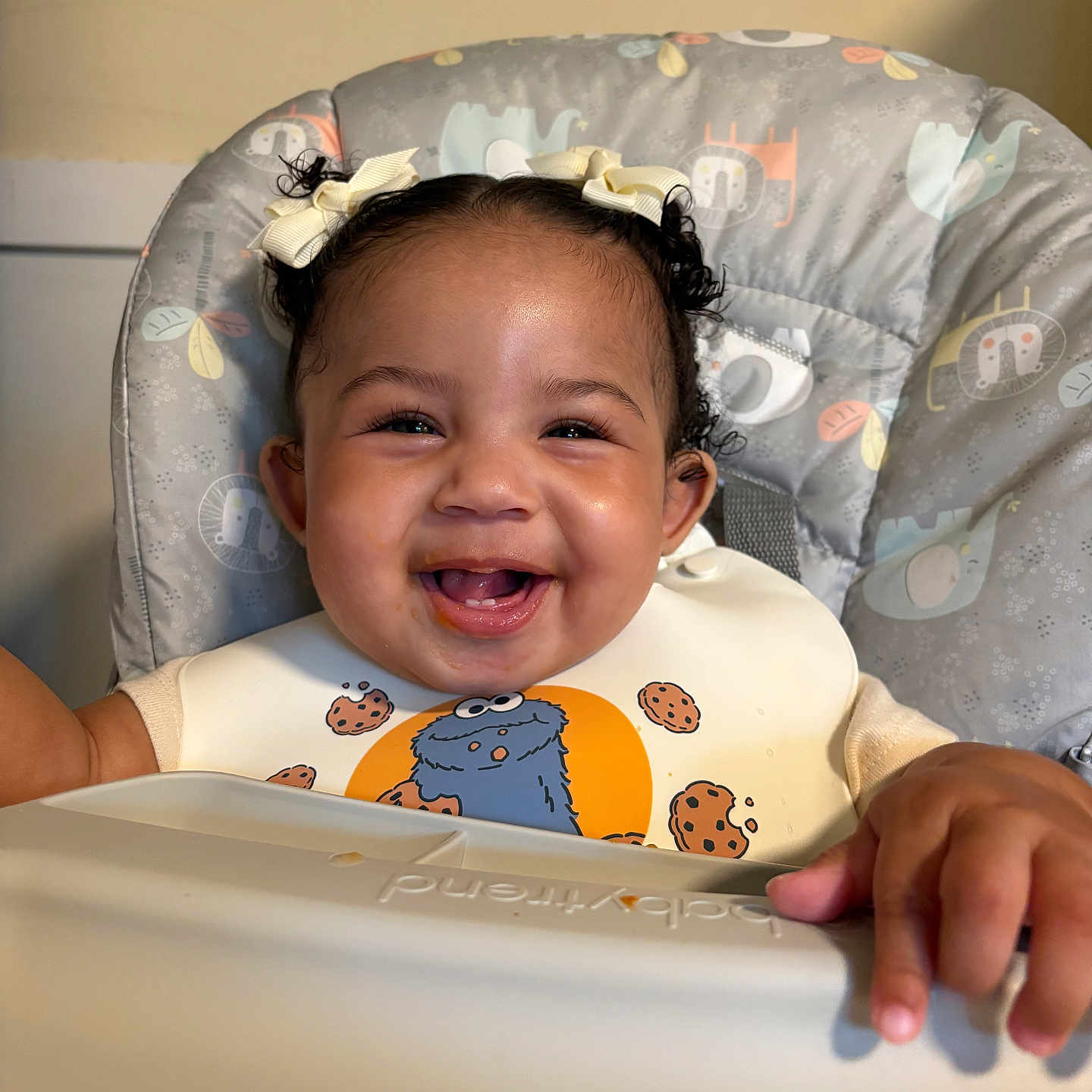 Kayani is registered to the contest to win money with this photo: baby, smiling, high_chair, bib, pigtails, bows, indoor, child, happy, face, person, clothing, seat, cute, infant, young_child, patterned, feeding, portrait, baby_food