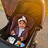 baby, stroller, child, outdoor, sunlight, smile, headband, curly_hair, seatbelt, toy, happy, daylight, path, infant, cute, summer, playful, sitting, person, accessory
