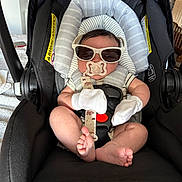 Malachi is registered to the contest to win money with this photo: baby, car_seat, sunglasses, pacifier, mittens, infant, child, seatbelt, striped_clothing, black_car_seat, indoor, cute, small_feet, relaxing, casual, comfort, safety_harness, head_support, adorable, portrait