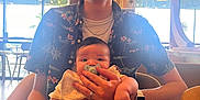 Malachi is registered to the contest to win money with this photo: adult, baby, pacifier, smiling, holding, indoor, cafe, table, chair, window, plant, natural_light, watch, ring, shirt, happy, person, family, casual, seated