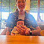 adult, baby, pacifier, smiling, holding, indoor, cafe, table, chair, window, plant, natural_light, watch, ring, shirt, happy, person, family, casual, seated