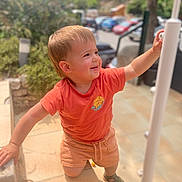 Jean a rejoint le concours — aidez-le/la à gagner de superbes lots ! casual, child, climbing, curly_hair, daytime, exploring, greenery, happy, orange_shirt, outdoor, person, playful, sandals, shorts, side_profile, smiling, stairs, summer_clothing, sunlight, toddler