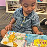child, denim_clothing, reading, picture_book, carpet, colorful, classroom, sitting, indoor, learning, preschool, kid, floor, shelf, toy_storage, person, young_child, engaged, education, casual_clothing