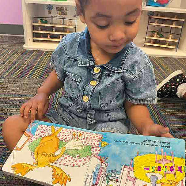 Jazelle is registered to the contest to win money with this photo: child, denim_clothing, reading, picture_book, carpet, colorful, classroom, sitting, indoor, learning, preschool, kid, floor, shelf, toy_storage, person, young_child, engaged, education, casual_clothing