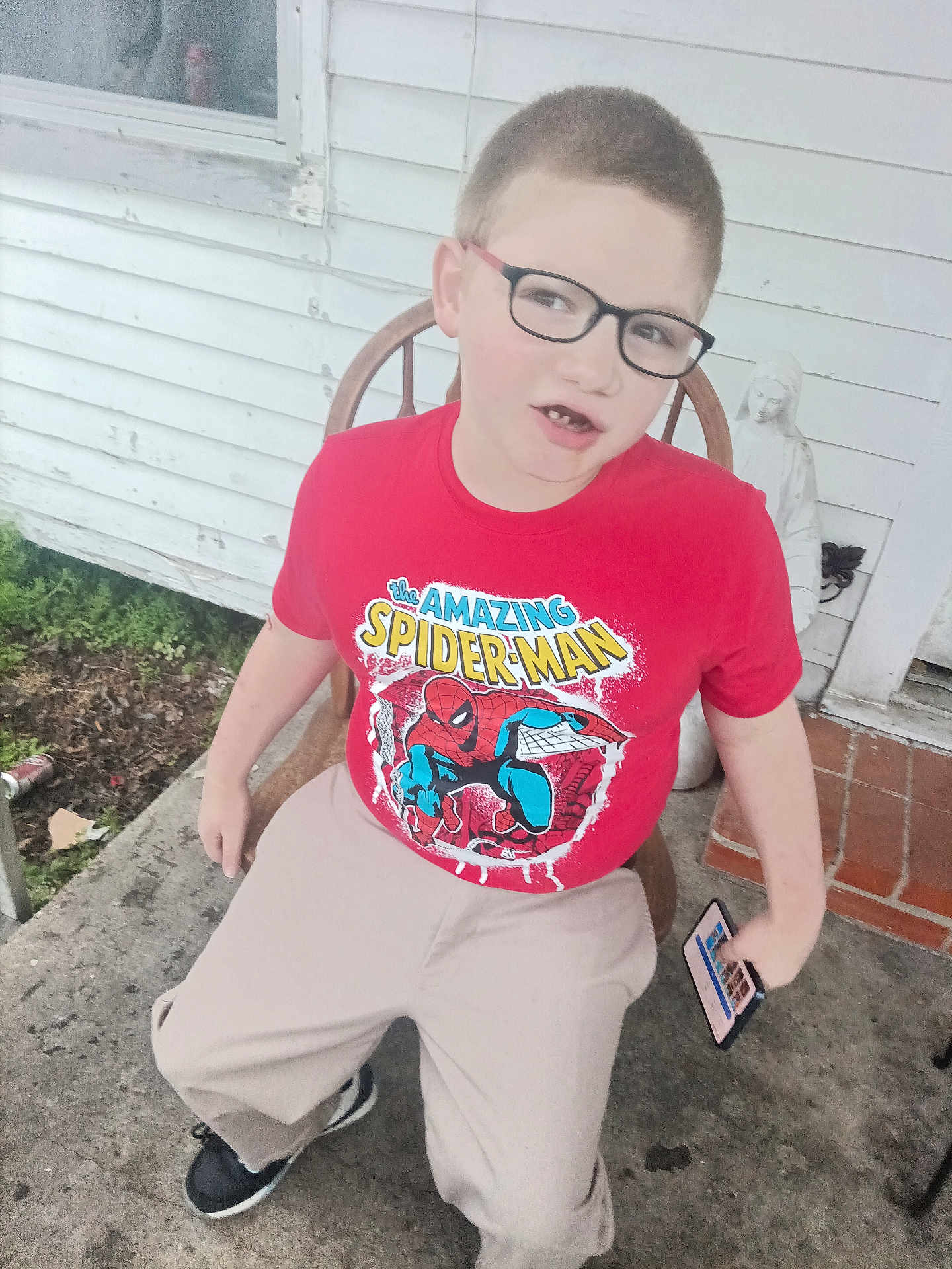 Karter is registered to the contest to win money with this photo: child, boy, glasses, red_shirt, spiderman, t_shirt, sitting, porch, chair, smartphone, concrete, house_siding, statue, missing_tooth, sneakers, casual_clothing, window, outdoor, smile, young_person