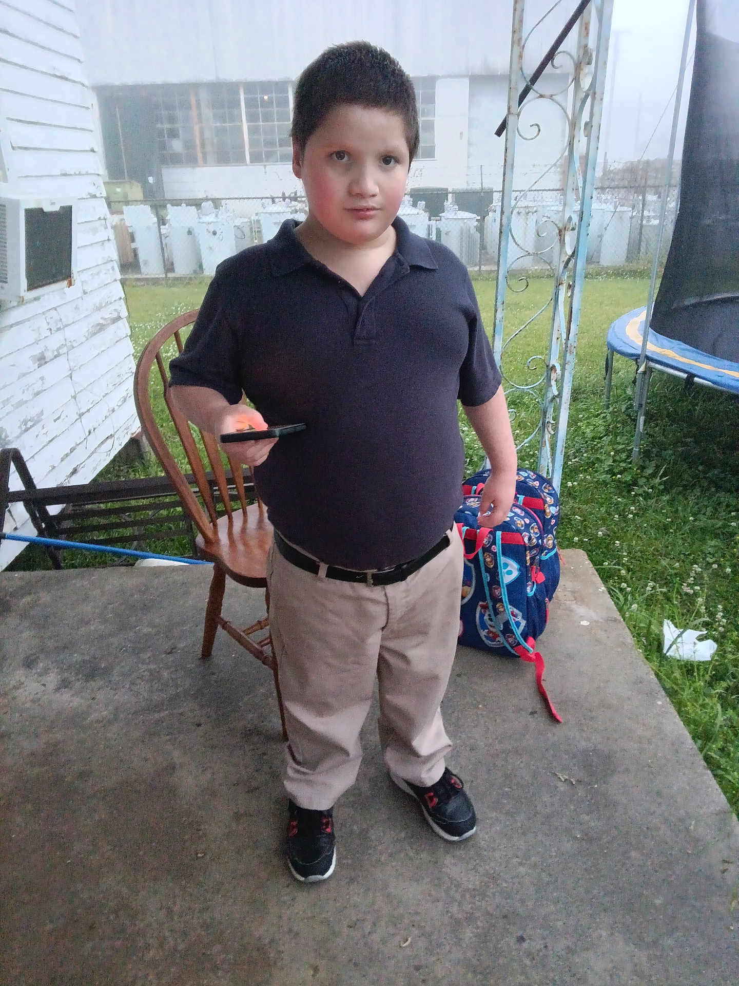 Karson joined the competition — help win amazing prizes! child, boy, standing, porch, backpack, smartphone, chair, polo_shirt, khaki_pants, sneakers, concrete, grass, trampoline, fence, building, outdoor, daytime, expression_neutral, short_hair, casual_clothes