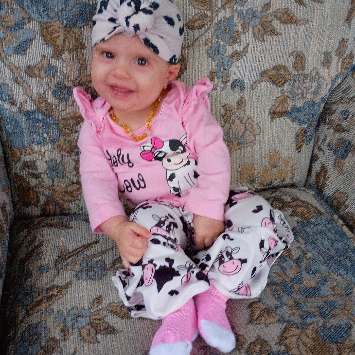 Myla is registered to the contest to win money with this photo: baby, child, smile, pink_clothing, headwrap, couch, floral_pattern, cow_print, socks, seated, indoors, portrait, cute, happy, person, infant, fashion, patterned_fabric, home, casual