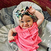 Chloe is registered to the contest to win money with this photo: baby, child, smiling, pink_clothing, headband, bow, raising_hand, blanket, indoor, cute, happy, infant, playful, portrait, person, fabric, pattern, skin, toddler, lying_down