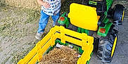 Wilder is registered to the contest to win money with this photo: child, boy, toy_tractor, hay, barn, cap, smiling, blue_jeans, shirt, floor, hay_bales, yellow, green, tractor, play, indoor, farm, vehicle, happy, clothing