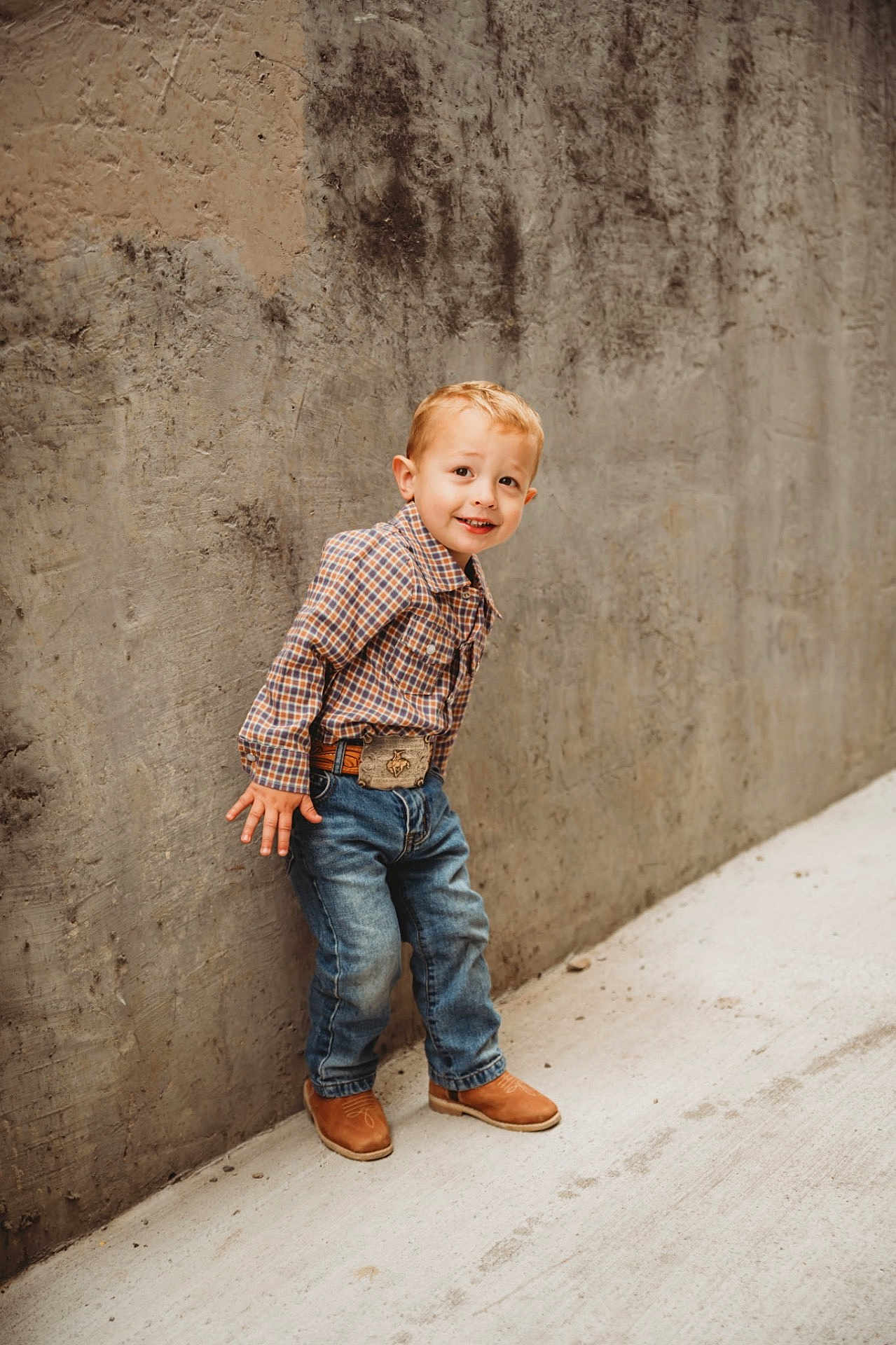 Wilder is registered to the contest to win money with this photo: child, boy, plaid_shirt, jeans, boots, belt, concrete_wall, sidewalk, smile, pose, short_hair, outdoor, casual_clothing, portrait, standing, young, fashion, urban, playful, cute