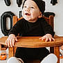 baby, child, smiling, high_chair, black_clothing, hat, wooden_furniture, toy_car, balloon, indoor, happy, face, hands, footwear, white_shoes, portrait, person, cute, seated, playful