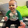 baby, child, smiling, dimples, sitting, shirt, green_sleeves, barefoot, indoor, pillow, blanket, checkered, happy, face, person, foot, hand, wall, carpet, cute