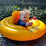 baby, inflatable_ring, water, pond, greenery, nature, smile, happy, child, outdoor, sunlight, grass, plant, floating, yellow, person, infant, summer, playful, water_plant