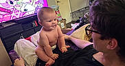 Theodore is registered to the contest to win money with this photo: baby, infant, child, adult, face, smiling, sitting, diaper, lap, family, indoor, living_room, television, cartoon, bed, blanket, pillow, pet_crate, home, portrait
