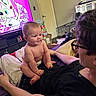 baby, infant, child, adult, face, smiling, sitting, diaper, lap, family, indoor, living_room, television, cartoon, bed, blanket, pillow, pet_crate, home, portrait