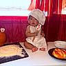 baby, child, chef_hat, spatula, flour, baking, bread, kitchen, curtains, table, plate, bowl, egg, utensils, pottery, red, white, cooking, apron, cute
