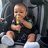 baby, child, car_seat, toy, socks, black_clothing, car_interior, seatbelt, cute, infant, hands, face, sitting, vehicle, window, headrest, footwear, person, indoor, colorful
