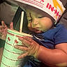 baby, child, cup, drink, straw, hand, hat, paper_hat, in_n_out, food_brand, blue_shirt, indoor, curious, close_up, face, portrait, young_child, seated, cute, chubby_cheeks
