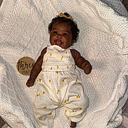 Zuri is registered to the contest to win money with this photo: baby, infant, smile, tongue_out, white_outfit, yellow_flowers, curly_hair, knit_blanket, texture, wooden_sign, three_months, lying_down, cute, happy, child, portrait, indoors, soft_lighting, closeup, person