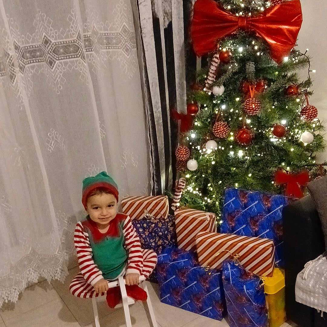 Jean-Joseph a rejoint le concours — aidez-le/la à gagner de superbes lots ! child, christmas_tree, cozy, curtains, elf_costume, festive, floor_tiles, giant_bow, gift_wrap, holiday_decor, indoor, ornaments, pajamas, presents, rocking_horse, smile, sofa, string_lights, toddler, wrapped_presents
