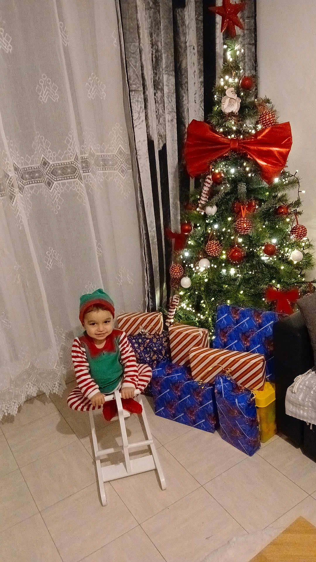 Jean-Joseph a rejoint le concours — aidez-le/la à gagner de superbes lots ! child, toddler, christmas_tree, presents, wrapped_presents, giant_bow, ornaments, string_lights, rocking_horse, pajamas, elf_costume, smile, curtains, floor_tiles, sofa, holiday_decor, indoor, festive, gift_wrap, cozy