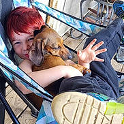 Twix is registered to the contest to win money with this photo: casual_clothing, chair, child, dachshund, dog, foot, friendship, hand, happy, hugging, metal_chair, outdoor, patterned_fabric, pet, red_hair, relaxed, resting, smiling, sneaker, wooden_floor