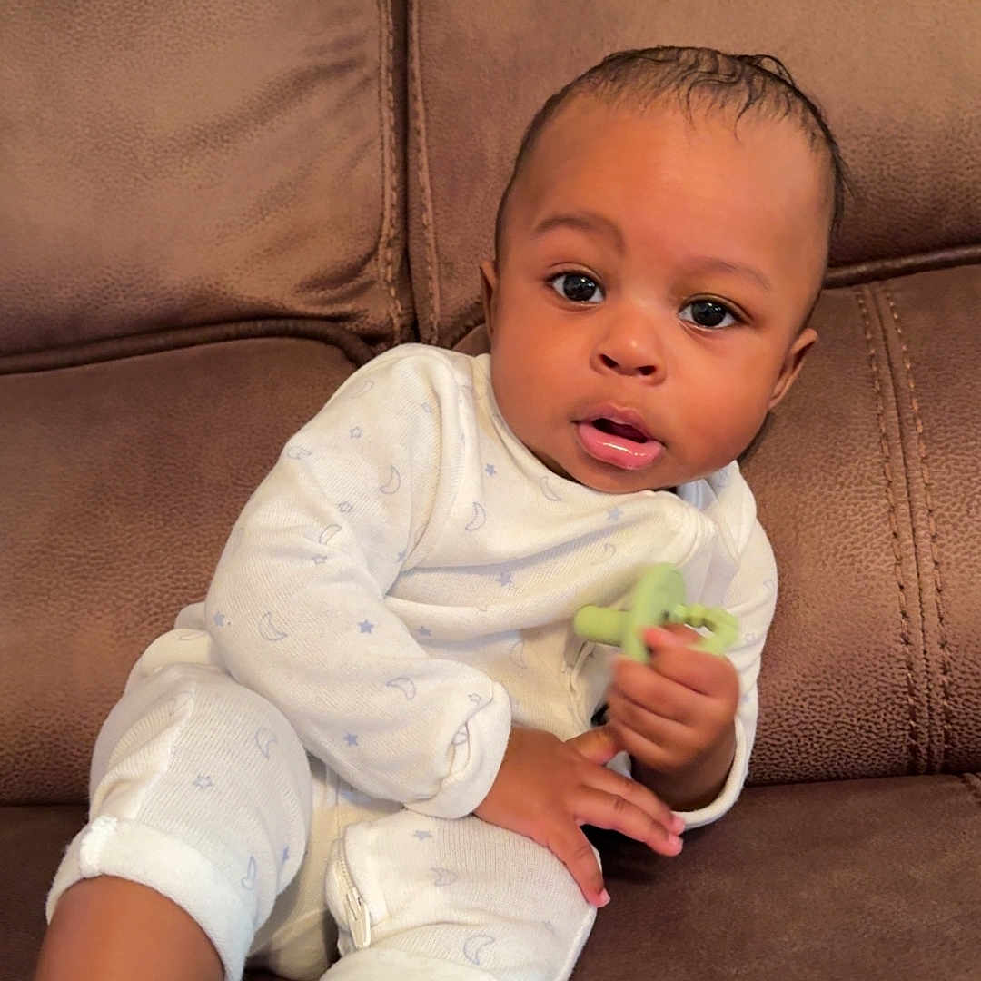 Taige is registered to the contest to win money with this photo: baby, black_socks, brown_furniture, casual, child, couch, curious_expression, cute, home, indoor, infant, portrait, relaxed, seated, small_hands, sofa, teething_toy, white_onesie, wide_eyes, young_child