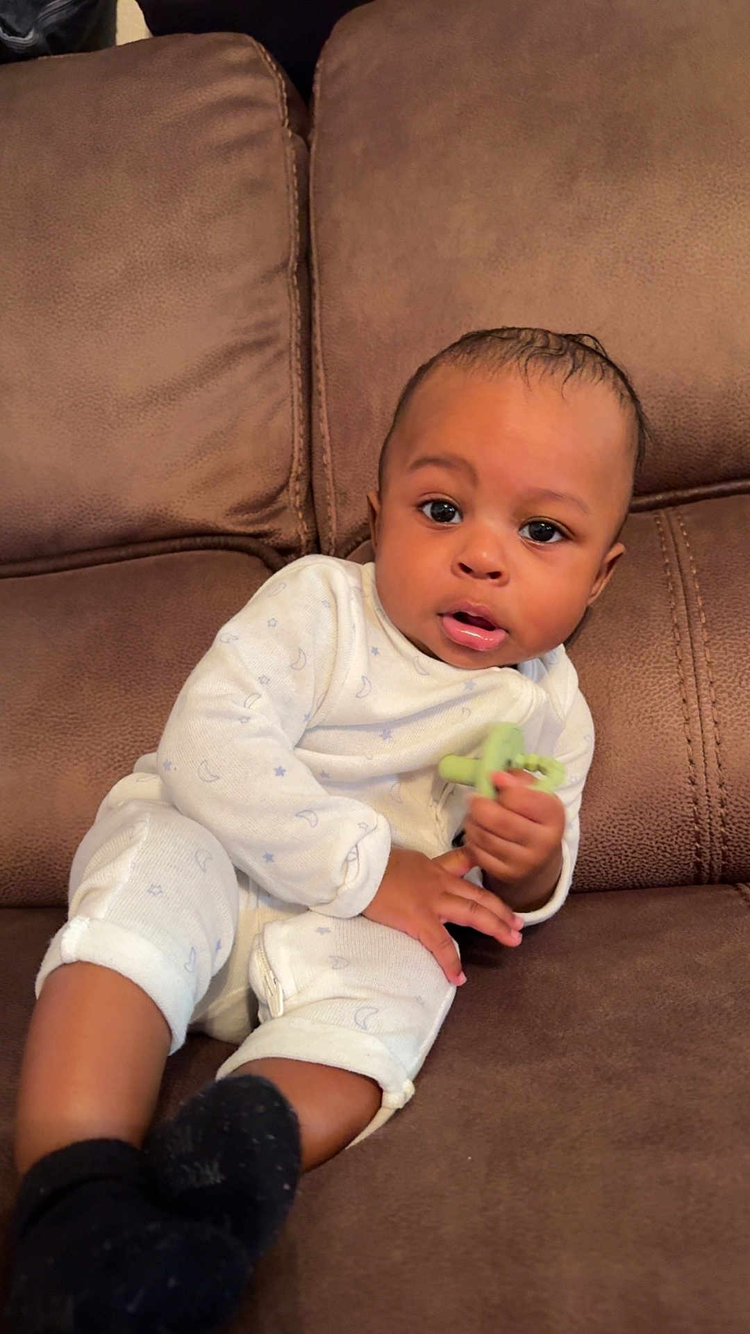 Taige is registered to the contest to win money with this photo: baby, child, infant, couch, sofa, white_onesie, black_socks, teething_toy, indoor, brown_furniture, curious_expression, seated, cute, small_hands, wide_eyes, relaxed, young_child, casual, home, portrait