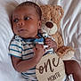 baby, bed, child, comfort, cozy, cute, face, hand, indoor, infant, lying_down, newborn, onesie, portrait, skin, soft_toys, striped_clothing, teddy_bear, white_bed, wooden_sign