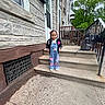 child, girl, stairs, backpack, building, sidewalk, railing, tree, outdoor, daylight, clothing, pink_bow, overalls, smiling, concrete, residential, urban, person, steps, casual